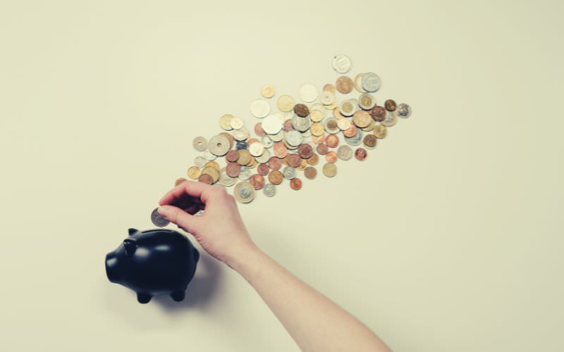A black piggy bank and many coins on a white background with a person putting a coin in it.